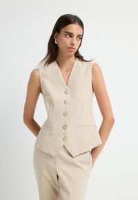 Gilet sans manches beige avec un col en V, design ajusté, fermeture à cinq boutons avec des accents brillants et deux poches avant. Texture de tissu lisse.