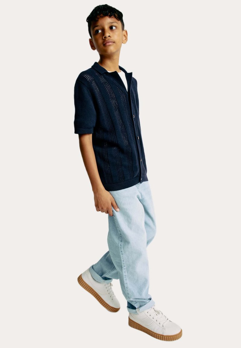 Cárdigan de punto azul marino de manga corta con detalles de tejido abierto, combinado con pantalones de mezclilla azul claro y zapatillas blancas con suelas texturizadas.