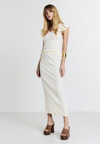 Haut blanc texturé avec des manches volantées, associé à une jupe longue et ajustée blanche. Sandales à plateforme marron avec orteils ouverts complètent le look.