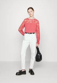 Top de manga longa coral com sobreposição de rendas, combinado com calças brancas e sapatos pretos. Asegura uma bolsa preta texturizada. Tecido suave, design ajustado.