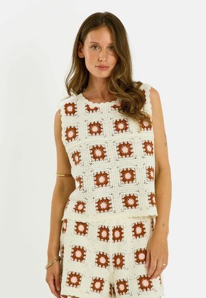 Femme portant un haut blanc sans manches en crochet et un short assorti avec des motifs carrés marron et rose, debout devant un fond blanc uni.