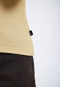 Waffle-textured, light yellow top with a rounded hem, featuring a black label. Paired with dark brown fabric below.