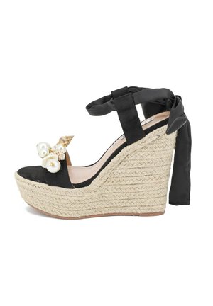 Sandale noire à talon compensé avec une plateforme en espadrille tissée texturée. Elle possède un lien à la cheville en ruban noir et des coquillages décoratifs avec des perles.