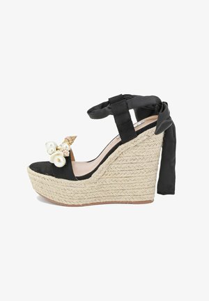 Sandale noire à talon compensé avec une plateforme en espadrille tissée texturée. Elle possède un lien à la cheville en ruban noir et des coquillages décoratifs avec des perles.