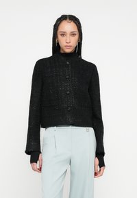 Jachetă neagră scurtă cu țesătură texturată, cu guler rotund, nasturi în față și mâneci lungi cu manșete tricotate negre.