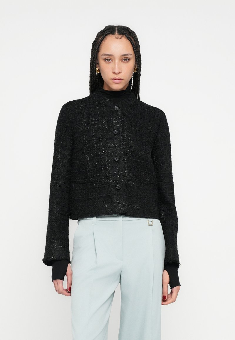 Jachetă neagră scurtă cu țesătură texturată, cu guler rotund, nasturi în față și mâneci lungi cu manșete tricotate negre.