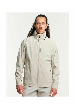 Man met krullend haar die een lichtbeige jas met rits en hoge kraag en bijpassende lichte broek draagt, staand tegen een effen achtergrond.