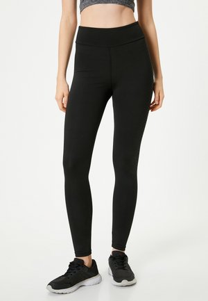 Zwarte leggings van rekbaar materiaal, met een hoge tailleband en textuurdetails langs de zijkant, gecombineerd met zwarte sportschoenen.