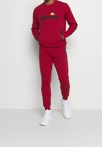 Survêtement rouge avec un logo "ellesse" noir sur le sweat à capuche. Comprend des poches zippées sur le pantalon. Porté avec des chaussures de sport blanches.