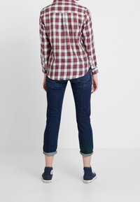 Camicia a quadri rossa e blu con maniche lunghe, abbinata a jeans scuri blu con risvolto e scarpe da ginnastica navy. La camicia presenta un dettaglio sul retro.