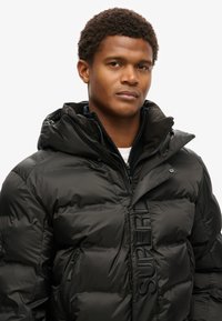 Svart pufferjacka med hög krage och dragkedjestängning. Har en strukturerad yta och ordet "SUPERDRY" präglat på framsidan.