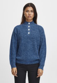 Pull bleu tricoté avec un col montant et quatre boutons blancs au niveau du cou, présentant un tissu texturé et une coupe décontractée.