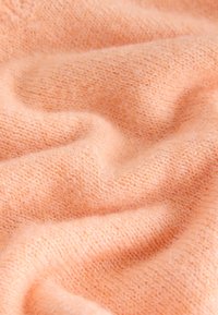 Miękka, brzoskwiniowa dzianina o teksturowanej, lekko puszystej powierzchni, ukazująca delikatne fale i średnią grubość zapewniającą ciepło.