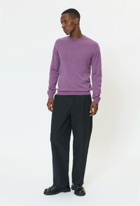 Vyras vilkintis violetinį ilgomis rankovėmis megztinį, juodus plačius kelnes ir juodus batelius su raišteliais, stovintis priešais paprastą baltą foną.