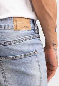 Lys blå denimjeans med en lærreim merket "REDEFINED REBEL" i livet. Har synlige sømmer og en baklomme.