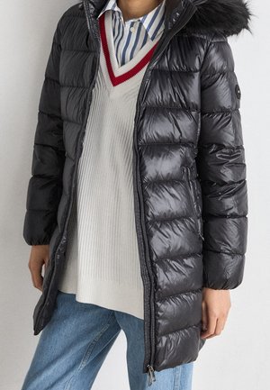 Personne portant un manteau matelassé noir avec capuche, un pull crème à liseré rouge, une chemise à rayures bleues et un jean bleu clair, debout à l'intérieur.