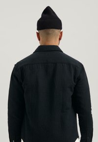 Camicia nera in tessuto a coste con colletto e maniche lunghe, caratterizzata da una cucitura visibile e un design uniforme. Berretto a coste nero sulla testa.