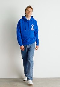 Sudadera azul con un bolsillo frontal y detalle gráfico blanco, combinada con jeans de color azul claro y zapatillas blancas, sobre un fondo neutro.