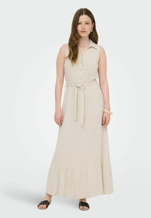 Nő hosszú barna hajjal, aki egy ujjatlan bézs, gombos maxi ruhát visel derékpánttal, fekete szandálokat és bézs karkötőt, sima háttér előtt áll.