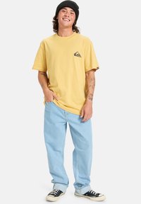 Žluté bavlněné tričko s logem na hrudi, v kombinaci se světle modrými volnými džíny a černými teniskami. Model nosí černou čepici.