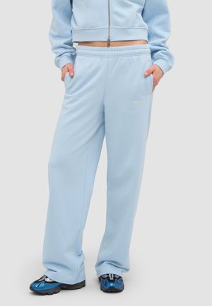 Person trägt hellblaue Jogginghose und passende Zip-up-Jacke mit den Händen in den Taschen, kombiniert mit blau-schwarzen Sneakers.