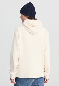 Felpa color crema con una superficie morbida, cappuccio con cordino e polsini a coste, abbinata a un berretto blu navy a coste, indossata di spalle.