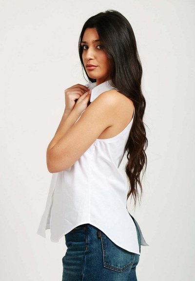Camicia bianca senza maniche con colletto e chiusura a bottoni, con orlo leggermente curvato, indossata con jeans in denim blu. Tessuto liscio, design minimale.