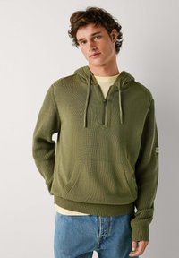 Sudadera de punto verde oliva con bolsillo frontal, cierre de media cremallera y capucha con cordón. Puños y dobladillo acanalados, llevada sobre una camisa de color amarillo claro.