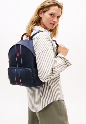 Marineblauwe rugzak met bruine leren accenten, voorzien van twee voorvakken en rood-wit gestreepte details. Soepele textuur, afgeronde vorm.