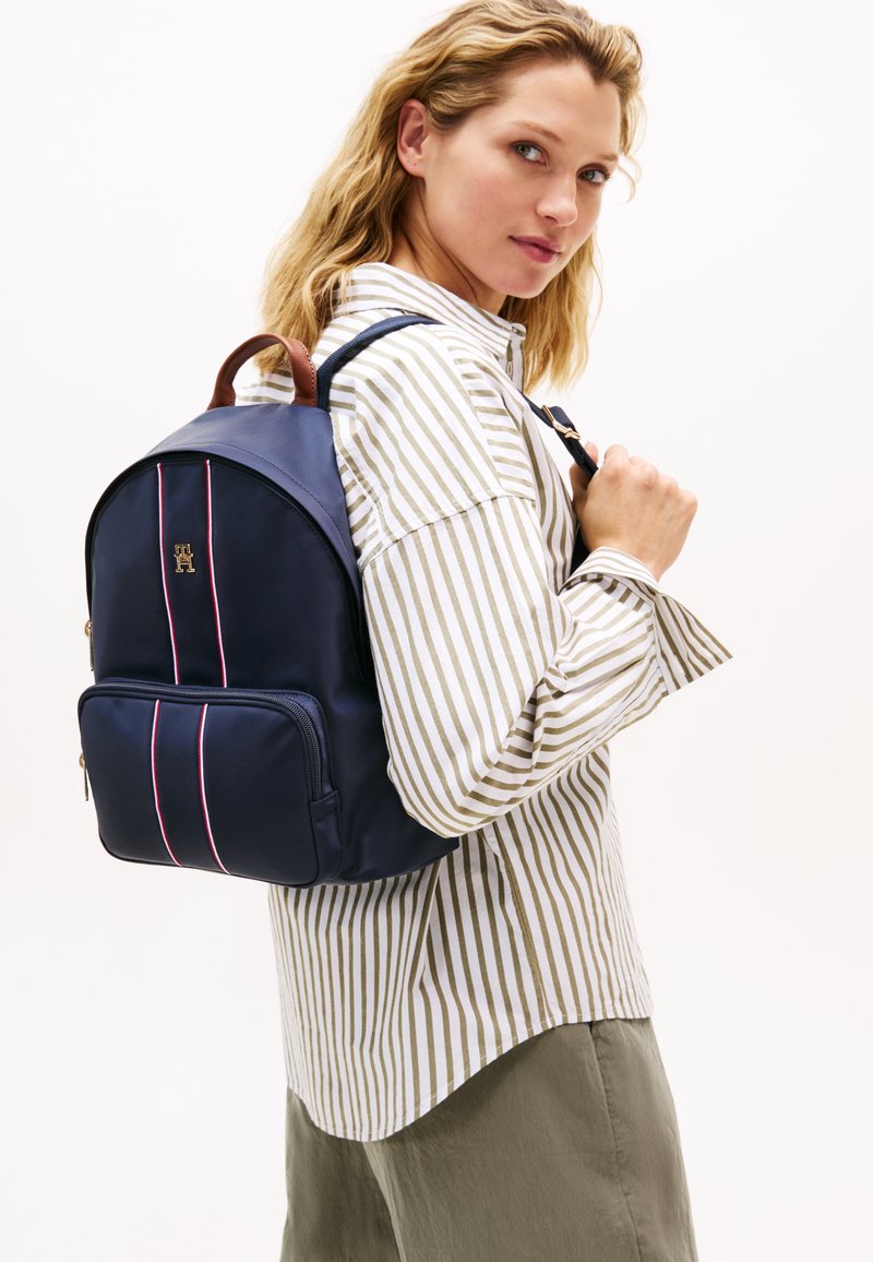 Σακίδιο πλάτης σε ναυτικό μπλε με καφέ δερμάτινες λεπτομέρειες, διαθέτει δύο μπροστινές τσέπες και κόκκινες και λευκές ριγέ λεπτομέρειες. Απαλή υφή, στρογγυλεμένο σχήμα.