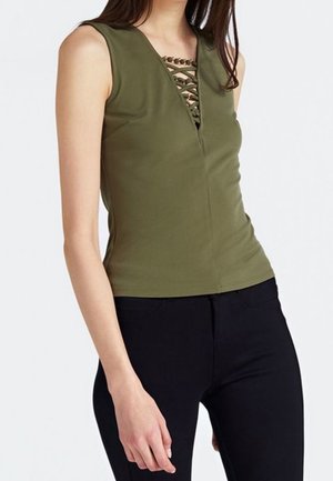 Vrouw draagt een mouwloze olijfgroene top met veterdetail aan de voorkant en zwarte nauwsluitende broek, staand tegen een witte achtergrond.