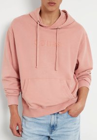 Sudadera de algodón rosa con capucha ajustable, bolsillo frontal y logo "GUESS" bordado. Cuenta con puños y dobladillo acanalados para un ajuste relajado.
