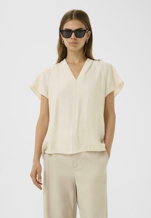Top beige de manga corta con escote en V, hecho de una tela ligera, con un corte relajado y costura visible en el centro.