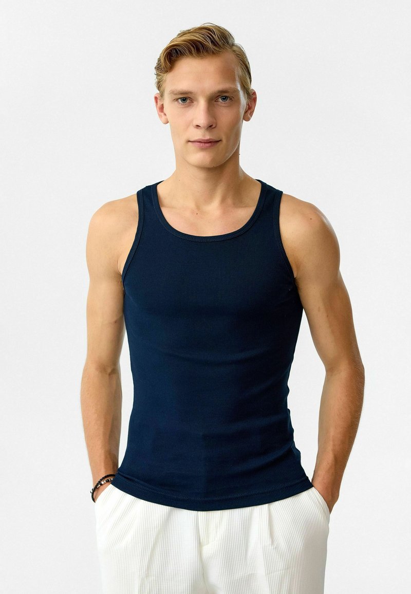 Marineblauwe geribde tanktop met een ronde halslijn, slim fit ontwerp en brede schouderbanden, gedragen onder lichtgekleurde broeken.