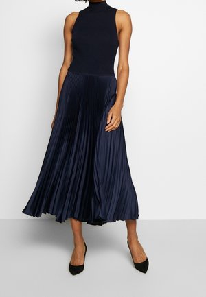 Kobieta w czarnym, bezrękawnym topie z golfem oraz granatowej plisowanej spódnicy midi, nosząca czarne szpilki z ostrym noskiem.