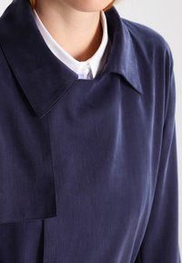 Námořnická modrá košile s velkým límcem a hladkou texturou, překrytá bílou košilí s límečkem, která má čistý design a minimální detaily.