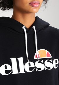 Sweat à capuche noir arborant un logo blanc proéminent avec un demi-cercle orange et rouge. Cordon de serrage blanc avec embouts argentés. Tissu doux et texturé.