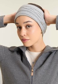 Fascia per la testa grigia in maglia con motivo testurizzato, caratterizzata da un piccolo logo metallico. Il modello indossa una giacca grigia con zip coordinata.
