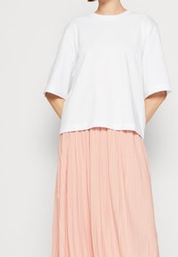 Valkoinen lyhythihainen t-paita pyöreällä pääntiellä, kerrostettuna vaaleanpunaisen laskostetun hameen päälle, joka korostaa pehmeää kangasta ja rentoa istuvuutta.