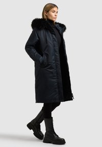 Lungo cappotto imbottito nero con cappuccio rifinito in pelliccia, tasche laterali e design elegante, abbinato a stivali da combattimento neri con lacci.