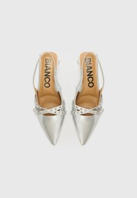 Hopeiset slingback-korkokengät, joissa on kärjet ja koristeellinen hihna sekä solki, valkoista taustaa vasten. Brändin nimi näkyy sisällä.