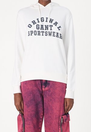 Felpa bianca con la scritta "ORIGINAL GANT SPORTSWEAR" stampata in blu navy. Presenta un cappuccio con cordino e polsini a coste. Abbinata a pantaloni cargo rosa, usurati.