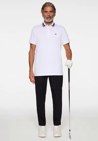 Valkoinen poolopaita, jossa mustaraidallinen kaulus, mustat housut ja valkoiset tennarit. Pitelee golfmailaa ja seisoo neutraalilla taustalla.