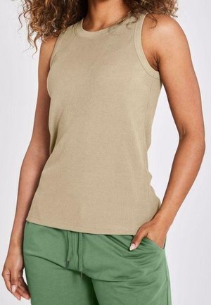 Vrouw met een beige mouwloze geribbelde top en groene broek met trekkoord, met één hand in haar zak tegen een effen achtergrond.