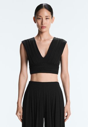 Kobieta z prostymi czarnymi włosami w ciasnym kokardce, ubrana w czarny krótki top bez rękawów oraz plisowane szerokie czarne spodnie.