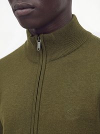 Gros plan d'une personne portant un pull zippé vert olive avec un col haut côtelé et un logo embossé sur la poitrine indiquant "EST 1999 VINTAGE."