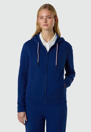 Giovane donna che indossa una felpa con cappuccio blu con cerniera e cordini rosa sopra una polo bianca, in piedi contro uno sfondo chiaro e uniforme.