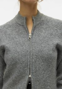 Suéter gris de punto con un cuello alto y una cremallera central. El material tiene un acabado suave y texturizado con un diseño ajustado.