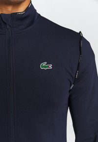 Marinblå zip-up jacka i stretchigt material, med en liten grön och r öd krokodil-logga samt LACOSTE-logotyp på kragen.