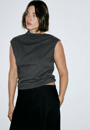 Femme aux cheveux courts et foncés portant un haut gris sans manches et un pantalon noir, debout avec une main dans la poche, regardant sur le côté.